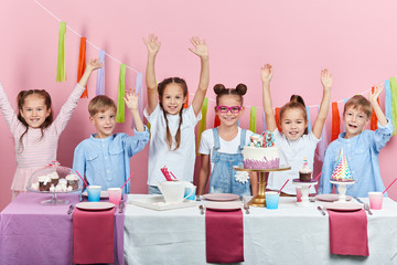 Group of six cheerful children standing at the table with cake on it holding hands up, holiday, festive time , isolated pink background