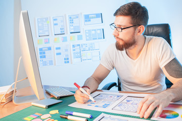 The designer develops the scheme of the mobile application. The man fills in the dialog box templates. Freelance.The young man looks at the computer screen and makes notes. Mobile app designer at work
