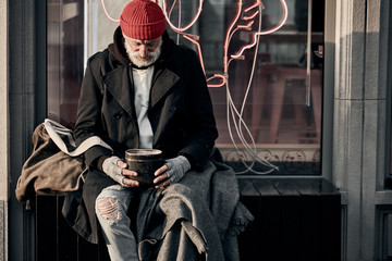 Mature caucasian vagrant man ask for some money donations from citizens, modern cafe behind him. Poverty, hungry, hope