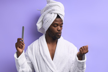 Cool handsome man in bathrobe doing manicure for himself with nail file isolated ober purple background