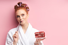 Young Redhead Woman Wearing Bathrobe, Curlers After Shower Eating Sweet Tasty Cake. Emotional Girl Posing With Cake Isolated Over Pink Background