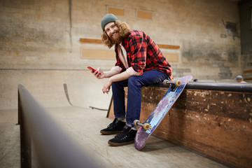 Wall Mural - Full length portrait of contemporary bearded man looking at camera and smiling while using smartphone siting on ramp in skating park, copy space