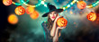 © Subbotina Anna - Halloween Witch holding Pumpkin lantern in a dark forest decorated with garlands and hanging Jack-o-Lantern Pumpkins. Beautiful young woman in witches hat and costume. Halloween party art design