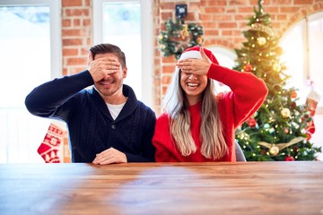 Poster - Young couple wearing santa claus hat sitting on chair and table around christmas tree at home smiling and laughing with hand on face covering eyes for surprise. Blind concept.