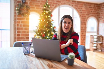 Canvas Print - Beautiful woman sitting at the table working with laptop at home around christmas tree winking looking at the camera with sexy expression, cheerful and happy face.