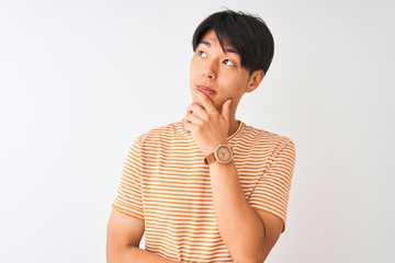 Wall Mural - Young chinese man wearing casual striped t-shirt standing over isolated white background with hand on chin thinking about question, pensive expression. Smiling with thoughtful face. Doubt concept.