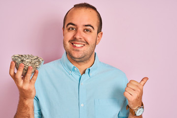 Canvas Print - Young man holding bowl with sunflowers seeds standing over isolated pink background pointing and showing with thumb up to the side with happy face smiling