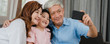 © tirachard - Asian grandparents selfie with granddaughter at home. Senior Chinese happy spend family time relax using mobile phone with young girl kid lying on sofa in living room. Panoramic banner background.