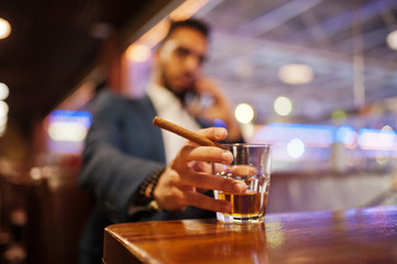 Wall Mural - Close up hand of handsome well-dressed arabian man with glass of whiskey and cigar posed at pub.