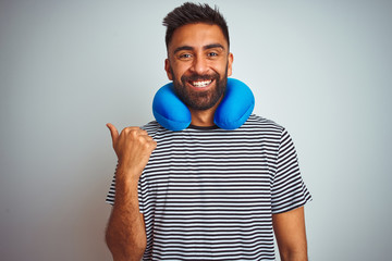 Young indian tourist man on travel wearing neckpillow over isolated white background smiling with happy face looking and pointing to the side with thumb up.