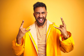 Wall Mural - Young indian man wearing raincoat standing over isolated yellow background shouting with crazy expression doing rock symbol with hands up. Music star. Heavy concept.