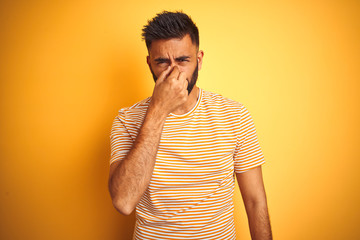 Canvas Print - Young indian man wearing t-shirt standing over isolated yellow background smelling something stinky and disgusting, intolerable smell, holding breath with fingers on nose. Bad smells concept.