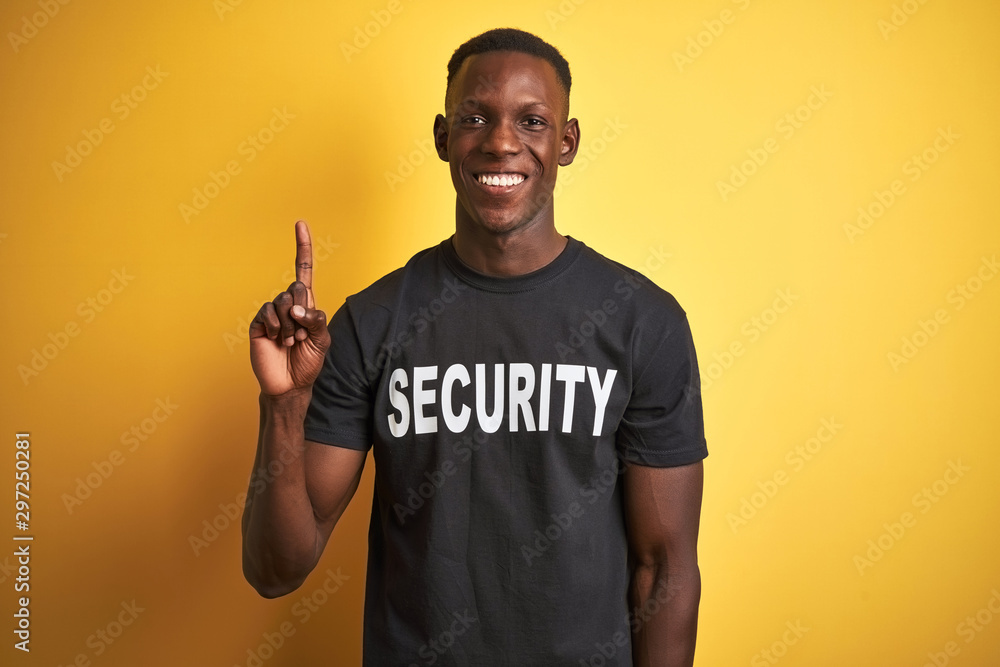 African american safeguard man wearing security uniform over isolated yellow background showing and pointing up with finger number one while smiling confident and happy. - obrazy, fototapety, plakaty 