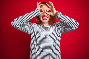 Wall Mural - Young redhead woman wearing strapes navy shirt standing over red isolated background doing ok gesture like binoculars sticking tongue out, eyes looking through fingers. Crazy expression.