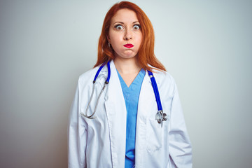 Canvas Print - Young redhead doctor woman using stethoscope over white isolated background puffing cheeks with funny face. Mouth inflated with air, crazy expression.