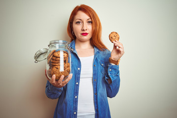 Sticker - Young beautiful redhead woman holding jar of cookies over white isolated background with a confident expression on smart face thinking serious