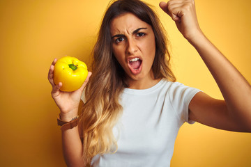 Poster - Young beautiful woman holding pepper over yellow isolated background annoyed and frustrated shouting with anger, crazy and yelling with raised hand, anger concept