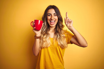 Sticker - Young beautiful woman holding  red cup of coffee over yellow isolated background surprised with an idea or question pointing finger with happy face, number one