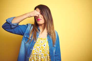 Sticker - Young beautiful woman standing over yellow isolated background smelling something stinky and disgusting, intolerable smell, holding breath with fingers on nose. Bad smells concept.