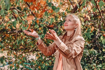 Beautiful young blonde girl throws leaves in the fall in the park. Happy smiling woman outdoors. Autumn concept.