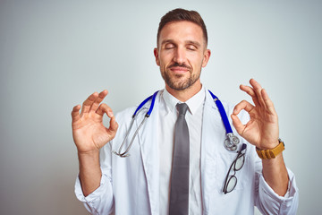 Poster - Young handsome doctor man wearing white professional coat over isolated background relax and smiling with eyes closed doing meditation gesture with fingers. Yoga concept.