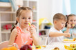 Cute child girl eats healthy food with kids group in kindergarten