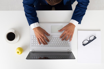 young businessman working in his computer in a desktop. He is wearing a blue jacket suit and a white shirt. On the desk there are a cup of coffee, an apple, his glasses and some documents