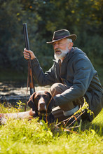Senior Good-looking Hunter With His Tracker Dog In Hunt On Wild Ducks. Sitting Near Lake, Wearing Hat, Holding Gun