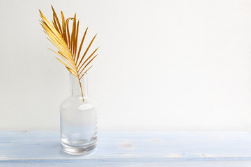 Wall Mural - Golden palm leaf in glass vase bottle on light blue wooden background. Creative minimal modern concept of organic beauty trend.