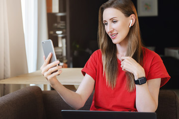 Wall Mural - Female looking at screen of smartphone, selecting songs for playlist and listening to music using wireless earphones. Young woman watching webinar on mobile phone. Girl using digital content for work.