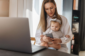 Wall Mural - Young woman texting on smartphone with baby girl sitting on her knees and looking at screen. Mother working at home distantly using wireless connected devices for job. Freelance vacancies for parents