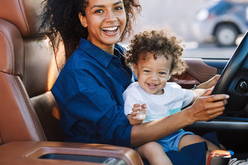 smiling mother with happy son looking at camera while sitting on car driver seat