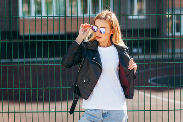 Wall Mural - Girl wearing t-shirt, glasses and leather jacket posing against street , urban clothing style. Street photography