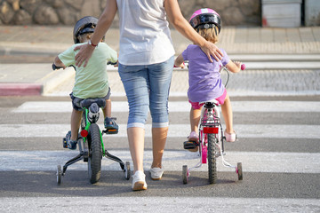 Mother goes pedestrian crossing with children on bicycles. A woman with children crossing the road in the city. Back view.     