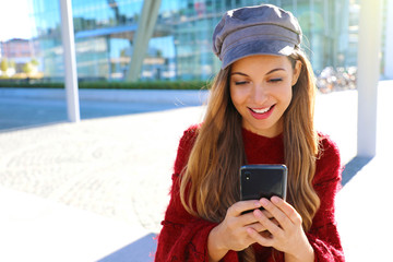 Wall Mural - Happy beautiful girl using smart phone in modern city. Fashion autumn winter girl portrait.