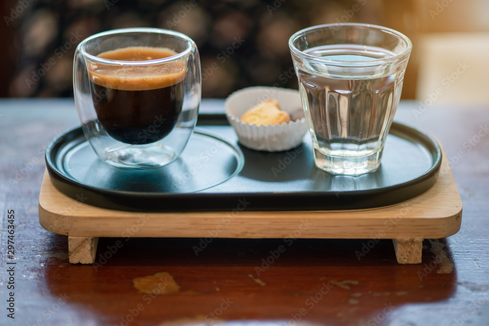 Hot espresso coffee in a clear glass, placed on a metal tray and wooden layer beside the coffee cup with a bear shaped cookie . All laid on dark brown wooden tables in retro coffee shop.