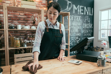 Wall Mural - Young asian chinese woman waitress in apron thoughtfully wiping counter at workplace in restaurant. female barista in white shirt cleaning in bright modern coffee shop. girl staff working in cafe