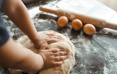 Small hands kneading dough. Little child preparing dough for backing. Kid's hands, some flour, wheat dough and rolling pin on the dark table.Cooking concept.