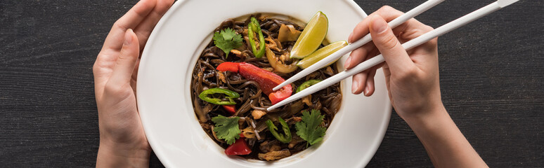 Wall Mural - cropped view of woman eating with chopsticks meat thai noodles on wooden grey surface, panoramic shot