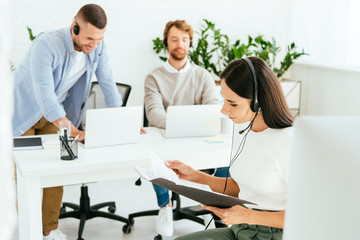 selective focus of attractive broker holding folder near coworkers using laptops in office