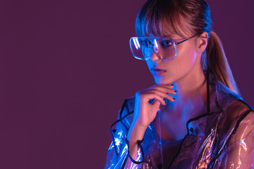 Wall Mural - Fashion sexy young woman girl model attractive face wearing stylish trendy transparent raincoat eyewear sunglasses looking away posing in neon light at purple magenta studio background copy space