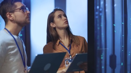 Young woman chief engineer talking with IT administrator solving system problems running maintenance diagnostics of computer server racks in modern data center.