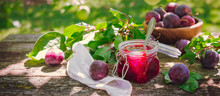 Glass Jar With Plum Jam Confiture And Ripe Plum Berries In A Basket On A Wooden Vintage Table In The Garden With A Copy Space, The Idea Of Home Canning And Organic Ecological Bio Nutrition
