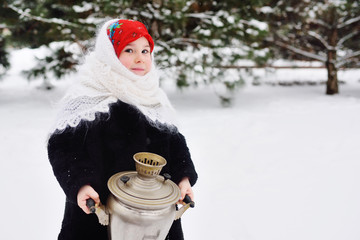 child girl in a fur coat and headscarf in Russian style holding a large samovar on the background of snow and forest.