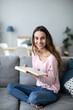 © BestForYou - Woman with a book on sofa looking up at the camera with a smile.