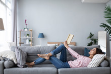 Young Beautiful Woman Is Reading A Book While Lying On A Sofa In The Living Room.