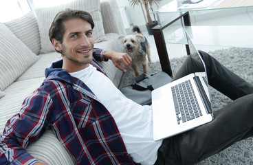 Wall Mural - smiling young man with laptop sitting on the floor near the couch