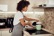 © nenadaksic - Mixed race woman in apron standing next to stove in domestic kitchen and making pasta and sauce.