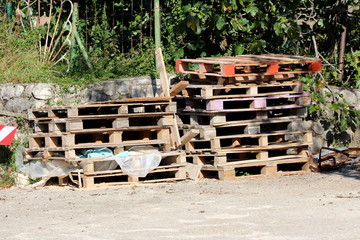 Wooden pallets stacked on two piles at local construction site in front of traditional stone wall surrounded with gravel and dense trees in background on warm sunny summer day