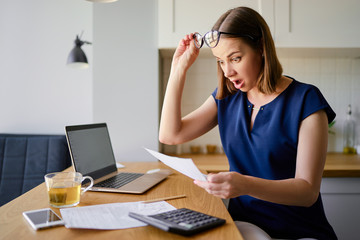 Stressed over bills. Portrait of surprised young woman using a laptop computer sitting at her kitchen holding utility bill and bank statements. Home interior.